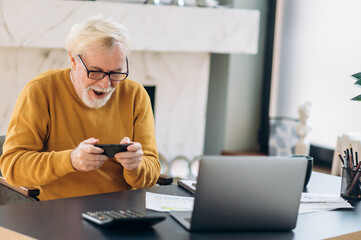 Cheerful mature gray-haired man emotionally playing a game on his phone during work break while sitting at his workplace © Kateryna
