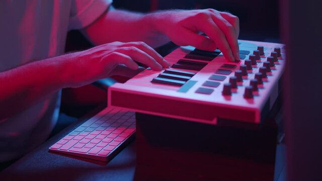 Male hands recording music, playing electronic keyboard, midi keys on the table with neon lights. Closeup of male hands composing music in night using midi controller