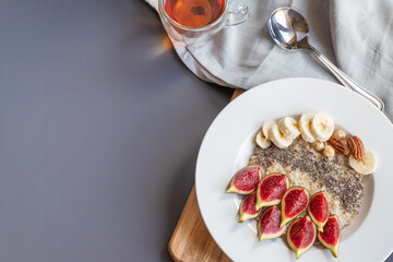 a bowl with breakfast oatmeal porridge with superfood chia seeds, fresh figs and banana on a wooden board on grey background , served with black tea, , shot from above