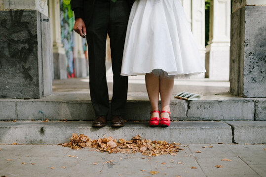 bride with bright red shoes with bow standing next to groom dressed in black with brow shoes