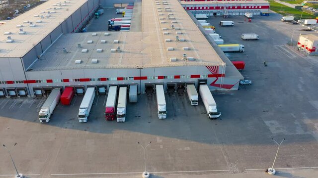 Logistics park with a warehouse - loading hub. Semi-trucks with freight trailers standing at the ramps for loading/unloading goods at sunset. Aerial hyper lapse (motion time lapse)