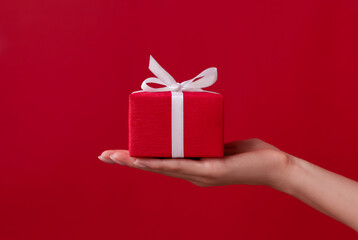 Female hand holding red gift box with white ribbon and bow on the red background. Christmas and New Year holidays. Valentine's Day and Birthday. Surprise, greeting, celebration, present. Isolated
