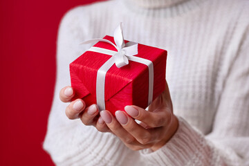 Female hands  holding red gift box with white ribbon and bow on the red background. Christmas and New Year holidays. Valentine's Day and Birthday. Surprise, greeting, celebration, present. Isolated