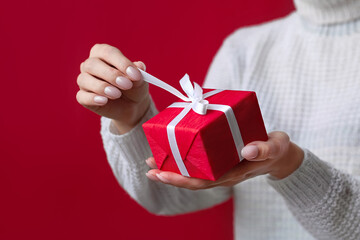 Female hands open Christmas red gift box with white bow on the red background. White sweater. New Year concept. Winter holidays and presents. Surprise delivery. Unpacking. Valentine's day, Birthday