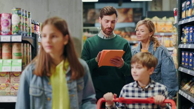 Happy family of four people walking through supermarket reading tablet checklist. Affectionate parents and kids shopping natural grocery foods in shop.