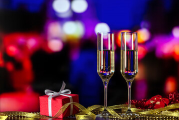 New Year's eve celebration. Two glasses with champagne with a gift and decorations against holiday lights. Toasting. Christmas background. Christmas card, greeting. Copy space. Blurred lights. Bokeh