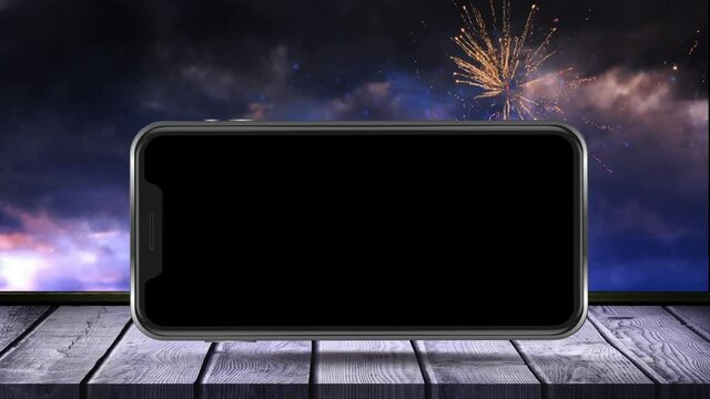 Animation of black smartphone screen on wooden surface with fireworks exploding in the background