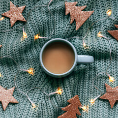A cup of coffee on knitted sweater with garland lights and Christmas decor, top view
