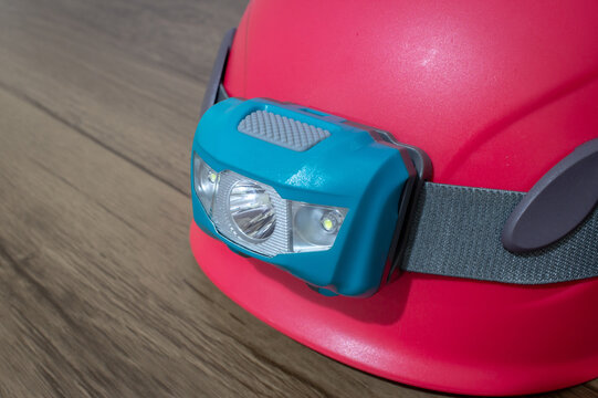 Teal Led Headlamp Turned Off Strapped To A Pink Climbing Helmet On A Wooden Table