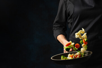 Professional chef in black uniform throws up frying mix of cabbage and carrot above the pan on dark blue background. Backstage of cooking meal. Frozen motion. Food banner concept.