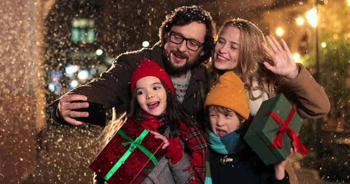 Cheerful Caucasian family taking funny pictures on smartphone together on snowy street on evening. Happy parents and kids taking selfie photos on cellphone in decorated xmas city