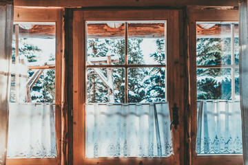 Bright Sunlight Shines Through the Wooden Cottage House Window. Beautiful snowy winter morning behind the chalet window. Sunshine and sparkling snow on fir trees in the cabin backyard