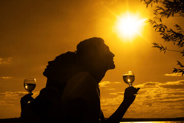 Silhouettes of Man and woman drink glasses of champagne wine at sunset dramatic yellow sky with clouds background Empty Copy space for inscription Couple against sun shine rays