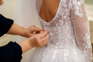 On the wedding day, the bridesmaid helps the bride to put on her wedding dress. The emphasis is on the hands fastening the buttons of the designer white dress. Preparing the bride to meet the groom