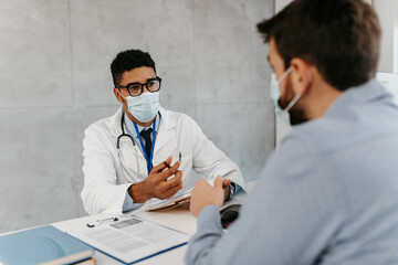 Male doctor wears protective face mask while talking with patient in clinic office during coronavirus. Pandemic, Covid-19 concept.