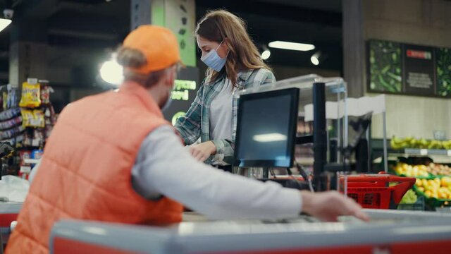 Young caucasian woman shopping in supermarket putting products on cash desk. Bearded male cashier worker scanning food items at cash register service. Grocery store.