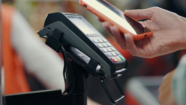 Close-up of female customer hand putting phone to terminal for contactless NFC payment in grocery store for goods. Shopping concept. Online transaction.