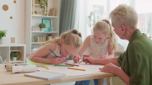 Medium over shoulder of cropper white-haired older woman sitting across desk of twin Caucasian granddaughters. Granny helping kids draw, family talking, smiling, having good time at home