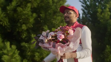 Waist up shot of male middle eastern courier in uniform and cap walking outdoors along the street with bouquet of flowers and document on clipboard