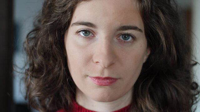 Close-up of beautiful confident young woman opening eyes and looking at camera. Attractive millennial girl with curly hair, red pullover and natural make-up at home indoors. Slow-motion.