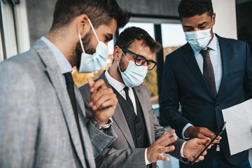 Group of businessmen working together in their office on important project. They are analyzing their business plan together. They are all wearing protective face masks because of Covid-19 pandemic.