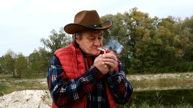 Mature adult man in cowboy hat and clothes, set fire cigarette from lighter and inhales smoke with pleasure. In countryside or ranch. Medium shot.