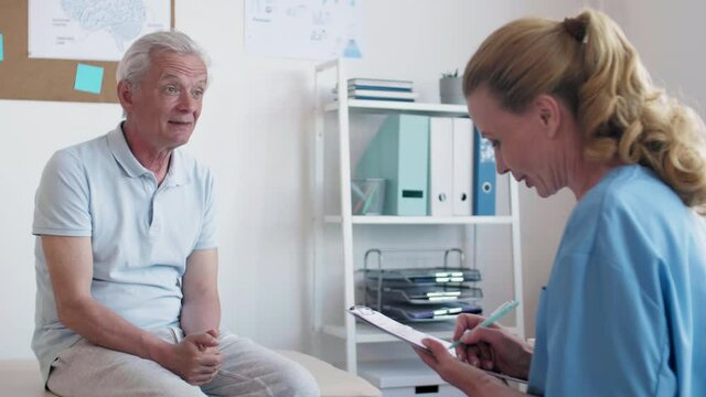 Rear view of Caucasian retired man sitting in medical office and communicating with female doctor. Doctor filling in patient card