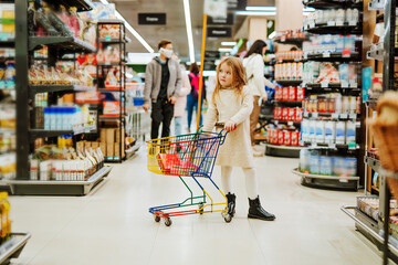 Little blonde girl l mask with trolley near shelves in supermarket