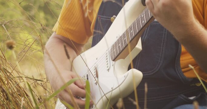 Musician's hands plays guitar as he sways in a grassy field during golden hour. (Close Up)