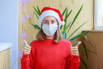 Obraz premium coronavirus, covid-19 in the new year. a woman in a red Santa hat and mask at home shows that everything will be fine