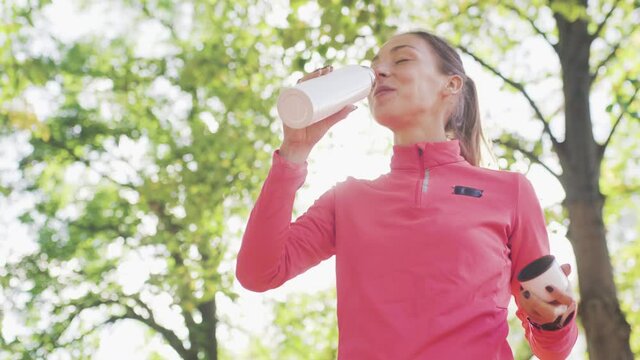 Fitness girl in pink sportswear drinking water from a bidon in the park