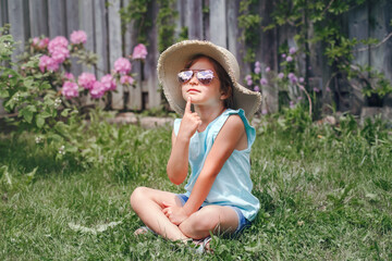 Portrait of cute adorable child girl in sunglasses and straw hat sitting on grass outdoors. Happy smiling Caucasian kid having fun at home backyard. Amazing joyful summer and lifestyle childhood.