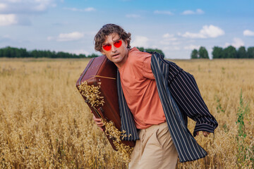 Tall handsome man with brown suitcase full of ears of oats standing at golden oat field.