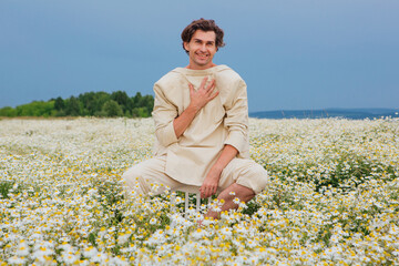 Fototapeta na wymiar Tall handsome man sitting on a back of a chair in camomile flowers field