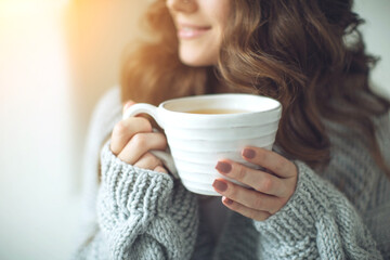 Close-up of female hands with a mug of drink. Beautiful girl in a gray sweater is holding a cup of...