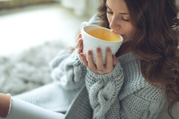 Close-up of female hands with a mug of drink. Beautiful girl in a gray sweater is holding a cup of tea or coffee in the morning sunlight. Mug for your design. High quality photo.