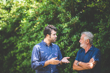 An adult hipster son and senior father talking at home, morning nature scene in the home garden outdoor