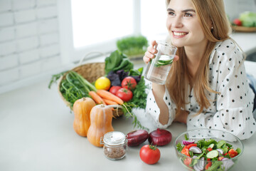 Young woman drinking water near the table with fruits and vegetables in the kitchen. High quality photo.