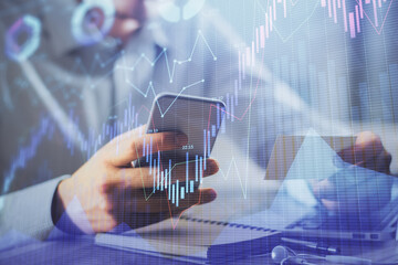 Double exposure of man's hands holding and using a phone and financial graph drawing. Analysis concept.