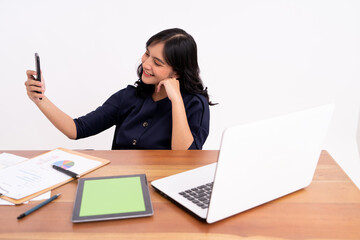 An attractive and cheerful business woman is taking a selfie and is smiling as she sits down. On modern office desk