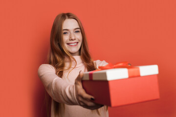 Fototapeta premium Adorable caucasian woman with freckles and red hair is giving at camera a present box smiling on a red studio wall