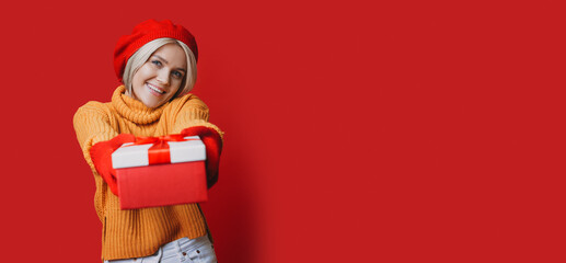 Caucasian woman with blonde hair and hat is receiving a present posing on a red wall at studio with free space