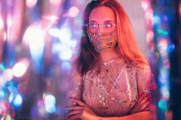 Christmas fashion look. Neon light portrait. New Year pandemic celebration. Glamorous woman in eyeglasses festive dress designer golden chain crystal beads face mask sparkle blur lens flare.