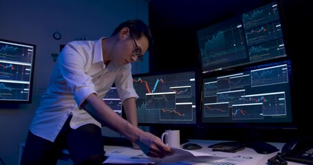 Asian business man is sitting on desk with monitors showing line graphs, charts. Alone male is drink coffee from cup while analyzing stock market trading, strategic planning of marketing at late night - Powered by Adobe