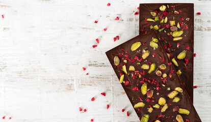 Fototapeta premium Two dark chocolate bars with sublimated raspberries and pistachios on white wooden background. Copy space. Top view. Horizontal orientation.