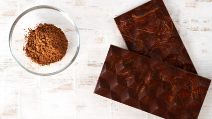Two homemade dark bitter chocolate bars with cocoa powder in glass small bawl on white wooden background. Bean to bar. Flat lay. Horizontal orientation.