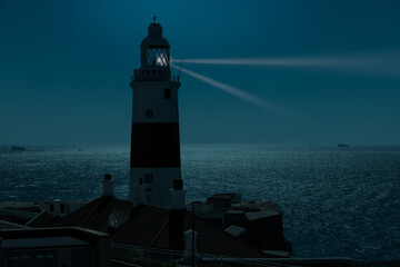 At night at the red and white trinity lighthouse on the British territory of Gibraltar. The moonlight illuminates the Strait of Gibraltar with some ships.
