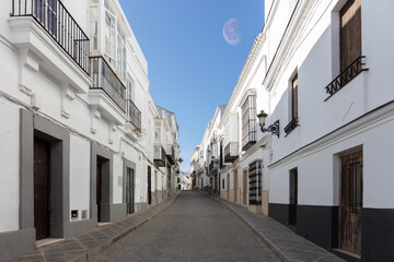 An alley in the historic mountain village of Medina-Sidonia in Andalusia in southern Spain. The houses are all painted white. It's early morning with sunshine. The moon is in the sky.