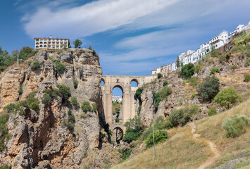 Fototapeta premium The famous arch bridge of Ronda in Andalusia in summer with a blue cloudy sky. The city is on top of the plateau. At the very bottom is a small waterfall.