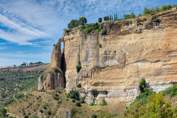Summer view of the high rock face of the Andalusian city of Ronda in sunshine and a blue sky with clouds. The rock on the left looks like a tower.